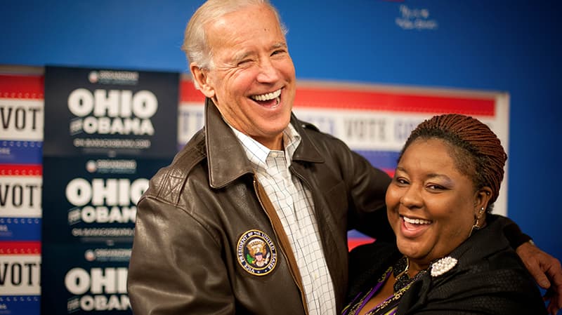 Joe Biden stands with his arm around a black woman while they are both laughing. He is wearing a brown leather jacket. Behind them on the wall are "Ohio for Obama" signs.