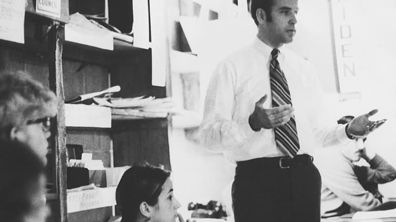 A black and white photo of Senator Joe Biden wearing a white button up shirt, standing with his arms raised beside him as he talks in an office.