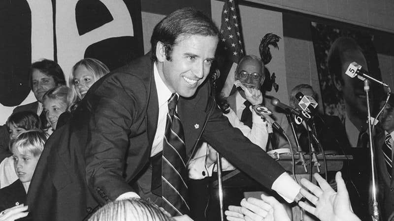 A black and white photo of Joe Biden standing on a stage while smiling. He is leaning down to touch the hands of those standing in the crowd.