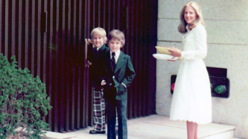 Young Jill Biden stands on on set of stairs wearing a white dress and holding a small bag. Her two sons are standing next to her and wearing suits.