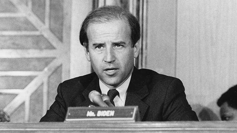 A black and white photo of then Senator Joe Biden sits behind a desk while speaking into a microphone. There is a nameplate in front of him that reads "Mr. Biden".