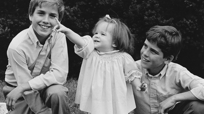 A black and white photo of Joe Biden's three children when they were young. Two boys kneel on either side of a young girl wearing a white dress and pointing towards the sky.