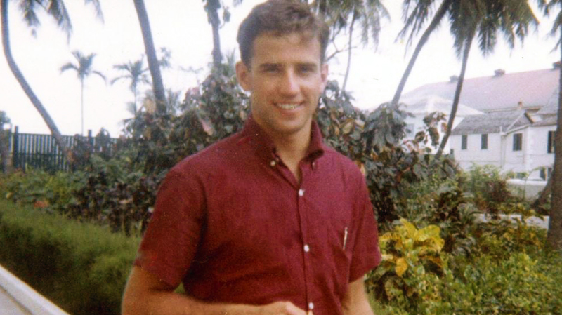 A young Joe Biden wearing a red shirt smiles while standing outdoors with palm trees behind him.