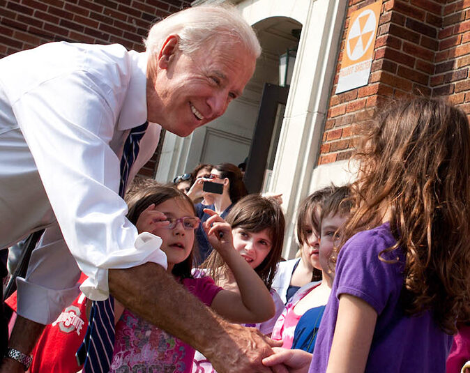 Joe Biden stands in a crowd of children while smiling and shaking hands with a young girl wearing a purple shirt.