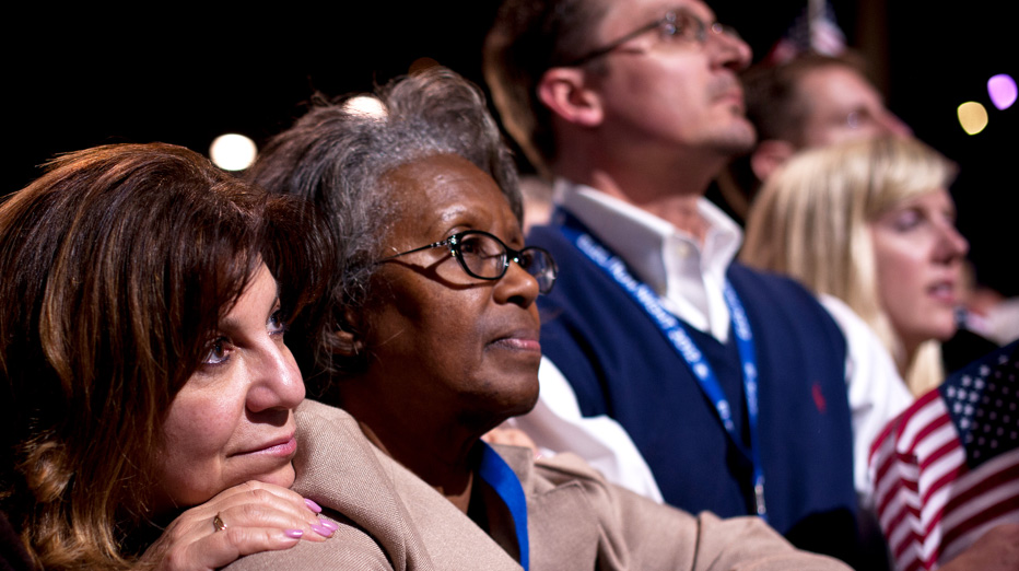 A white woman and an african american woman watch someone speak at a rally. The white woman is resting her head on the other's shoulder.