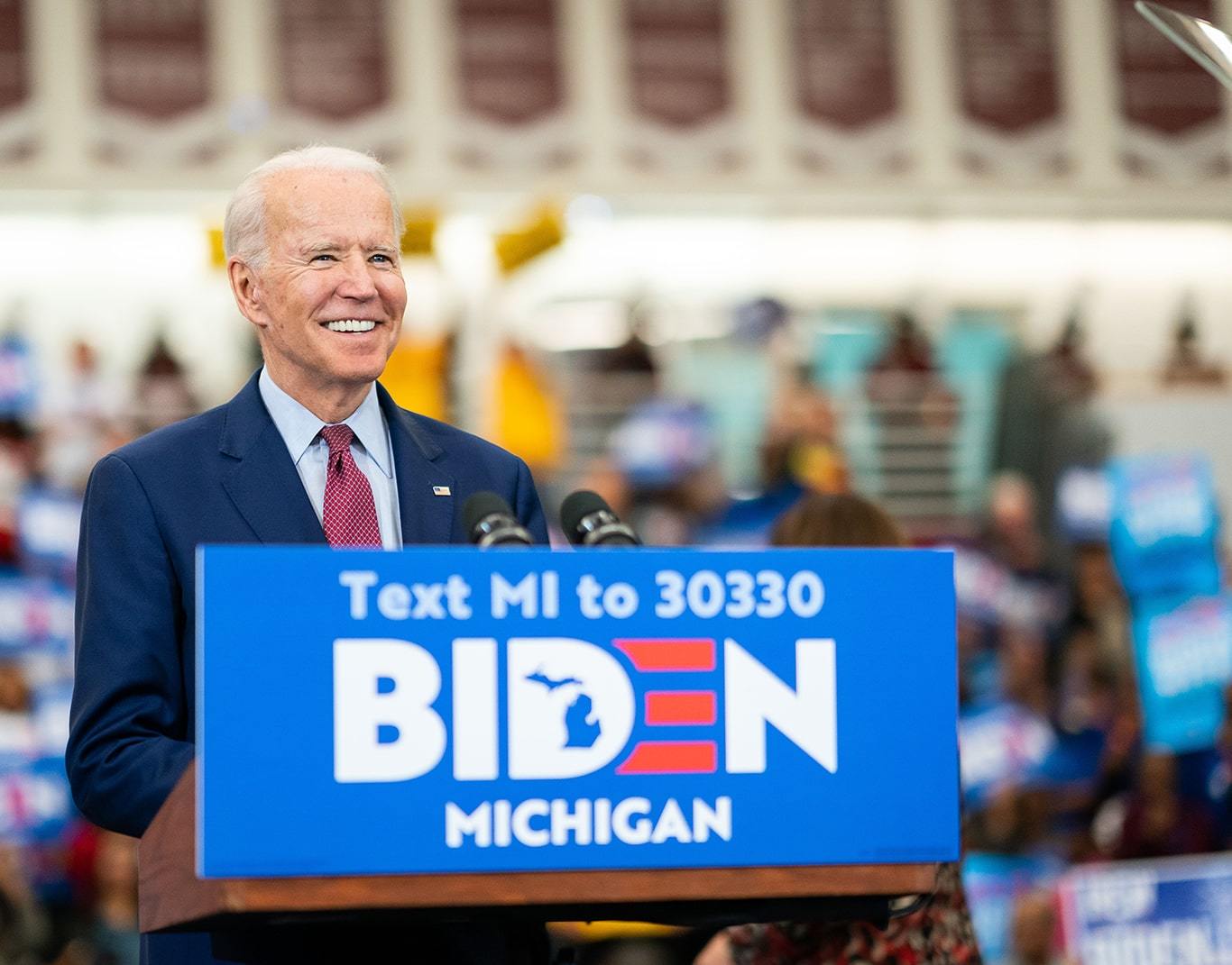 Joe Biden smiling at a podium during a rally in Michigan. He is wearing a blue suit, red tie, and an American flag pin.