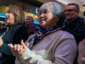 Two Asian American women standing up at a large gathering. They are smiling, clapping, and cheering.