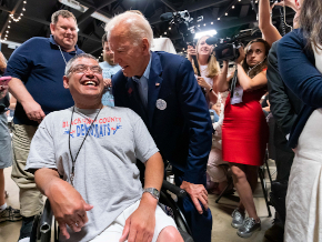A man in a wheel chair wearing a grey t-shirt smiling. Joe is next to him in a blue suit, and there is a crowd of people around them smiling and taking photos.