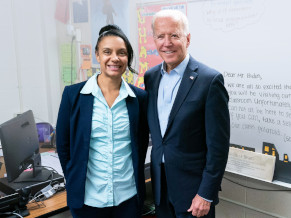 Joe in a suit with no tie, standing next to a female teacher wearing a light blue shirt and navy sweater. They are standing in front of a whiteboard in a classroom and smiling at the camera.
