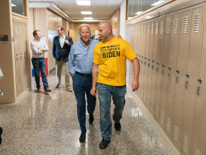 Joe, in a blue shirt and slacks, walking down a hallway of a school next to a man wearing a yellow Fire Fighters for Biden shirt. There are lockers on either side of the hallway and a couple other men in the background. Joe has his arm around the firefighter.