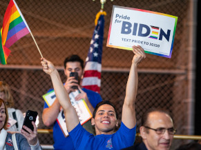 Close up of a young man holding up a rainbow flag in his right hand and a Pride for Biden sign in his left. He is smiling and appears to be in a crowd at an event or rally.