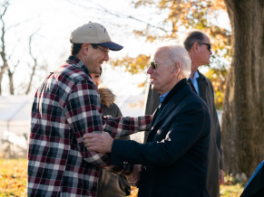 Joe shaking hands and grasping the arm of a supporter. Joe is wearing sunglasses and a coat and the man is wearing a hat and flannel shirt. They are outside and there are trees with leaves changing color in the background.