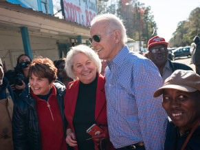 Joe outside wearing sunglasses and a blue checked shirt, surrounded by a small crowd of seniors. They are smiling and talking and not looking direclty at the camera.
