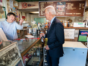 Joe wearing a suit in a small shop with a menu board in the background that reads Hanks Hoagies. Joe is talking to the business owner, who is wearing a light blue shirt and standing behind a counter.