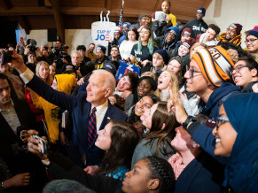 Joe wearing a suit surrounded by a crowd of college aged students. Joe is taking a selfie with the students who are leaning in and smiling at the camera.