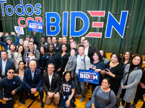 Joe kneeling with a crowd of Latino supporters. They are in front of a sign that says Todos Con Biden and are holding California for Biden signs. Everyone is smiling.